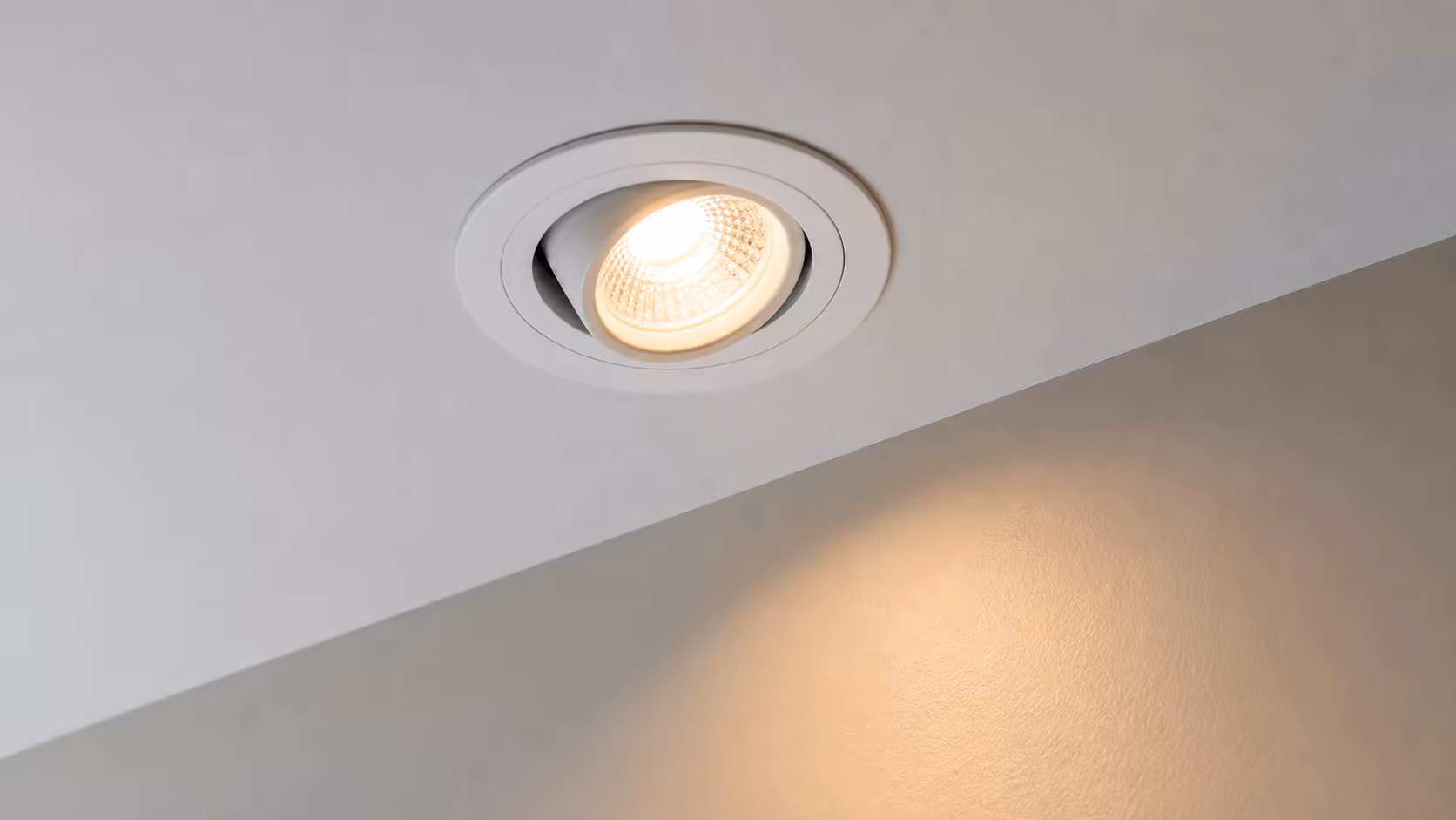 Détail en gros plan d'un spot LED encastré orientable fraîchement posé dans un faux plafond placo blanc mat, halo lumineux chaud sur le mur adjacent
