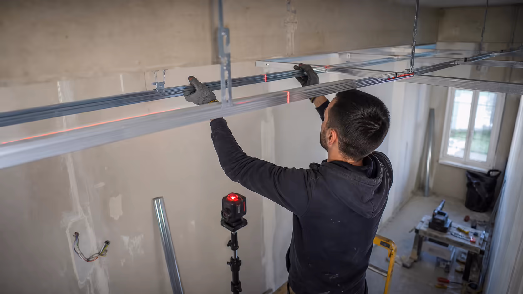 Plaquiste en cours de pose d'ossature métallique au plafond d'une pièce, niveau laser allumé, rails F47 alignés, photo en plongée